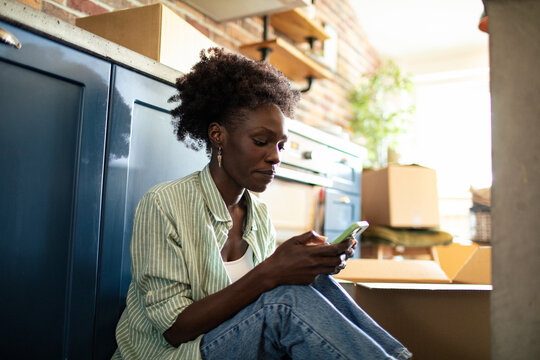 Young woman sitting on kitchen floor using smartphone after moving in
