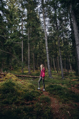 Naklejka na ściany i meble a woman in a pink jacket travels through the mountains. go on a hike with friends. a woman stands in the middle of a spruce forest
