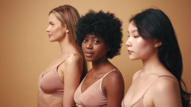 Multi-ethnic multiracial young women in beige lingerie isolated on a beige background. African-American, Caucasian and Asian women posing in the studio. Concept of face body skin care, cosmetology.
