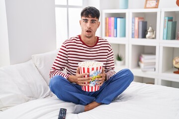 Young hispanic man eating popcorn sitting on the bed watching a movie in shock face, looking...
