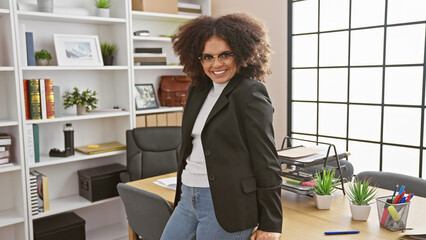 A young, attractive hispanic woman with curly hair smiling confidently in a modern office interior.