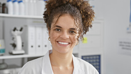 Smiling young hispanic woman with curly hair dressed in a lab coat standing in a medical laboratory