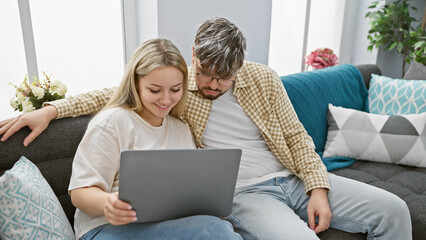A young man and woman sit closely on a sofa in a cozy living room, engaged in viewing a laptop screen together.
