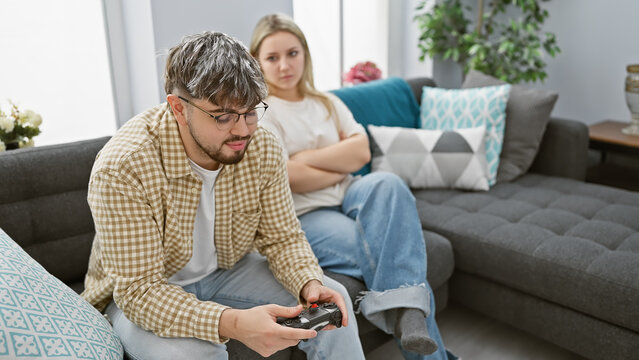 A woman looks on displeased as her partner focusedly plays video games in their cozy living room.