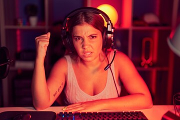 Young blonde woman playing video games wearing headphones angry and mad raising fist frustrated and...