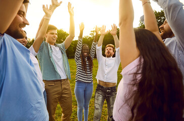 Portrait of a group of excited happy young friends students or colleagues having fun hanging together outdoors in the summer park. People standing in a circle raising their hands up enjoying meeting.
