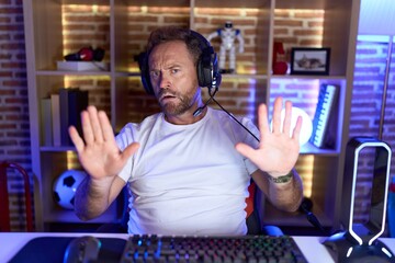 Middle age man with beard playing video games wearing headphones moving away hands palms showing...
