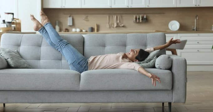 Cheerful carefree young Indian woman lying on back on comfortable couch, outstretching body, legs, opening hands, smiling with closed eyes, laughing, enjoying leisure, homey comfort