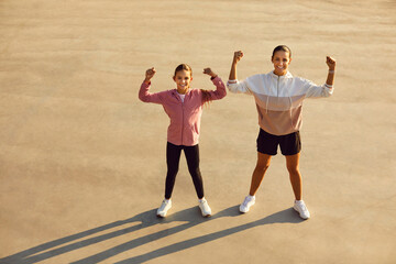 Happy sporty mother with child daughter in sportswear standing in the park outdoors with hands up in fist and looking at camera. Exercising in nature, family sport and healthy lifestyle concept