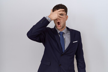 Young hispanic business man wearing suit and tie peeking in shock covering face and eyes with hand, looking through fingers with embarrassed expression.