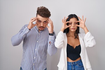 Young hispanic couple standing over white background trying to open eyes with fingers, sleepy and tired for morning fatigue