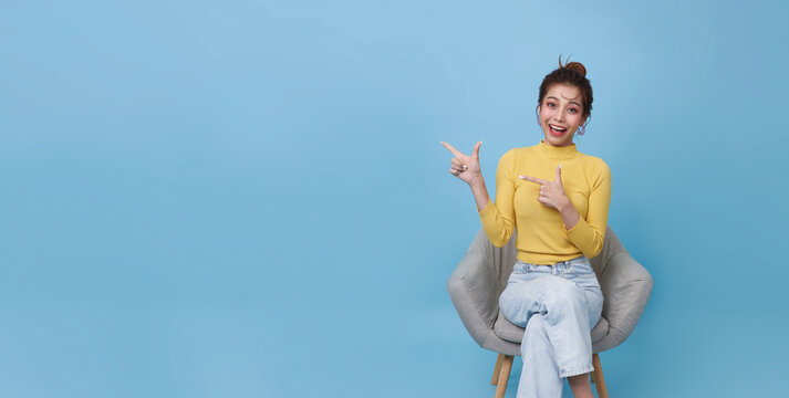 Excited young Asian woman sitting in armchair, pointing fingers offering space for your advertisement against blue studio wall. Best promotion, cool discount, huge sale concept