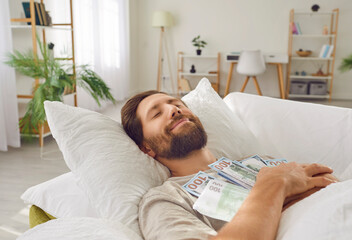 Smiling cheerful sleeping man lying in bed with closed eyes, embracing lots of cash dollar...