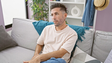 Handsome young hispanic man with a serious expression, sitting in comfort on a cozy sofa, relaxing and resting in his stylish indoor apartment living room at home