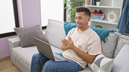 Confident young hispanic man enjoys relaxing on his comfortable sofa, using gadget at home while engaging in an exciting video call, comfortably sitting in the warm sunlight of his living room.