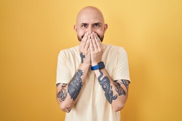 Hispanic man with tattoos standing over yellow background laughing and embarrassed giggle covering...