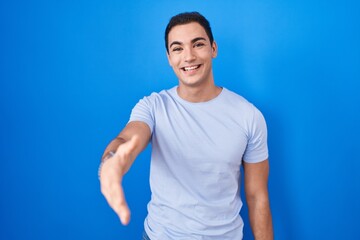 Young hispanic man standing over blue background smiling friendly offering handshake as greeting and welcoming. successful business.