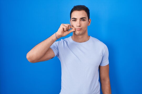 Young hispanic man standing over blue background mouth and lips shut as zip with fingers. secret and silent, taboo talking