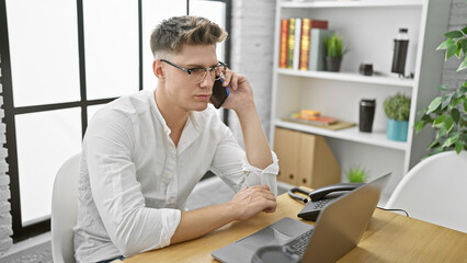 Fototapeta premium Attractive young caucasian man, focused business worker, talking on smartphone while working on laptop in office