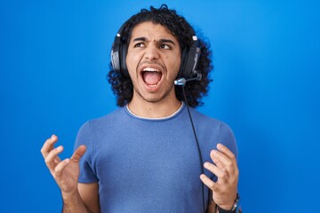 Hispanic man with curly hair listening to music using headphones crazy and mad shouting and yelling...