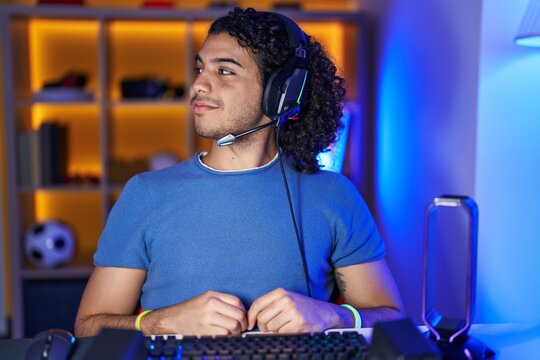 Hispanic Man With Curly Hair Playing Video Games Looking To Side, Relax Profile Pose With Natural Face And Confident Smile.