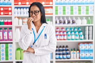 Hispanic woman working at pharmacy drugstore looking stressed and nervous with hands on mouth...