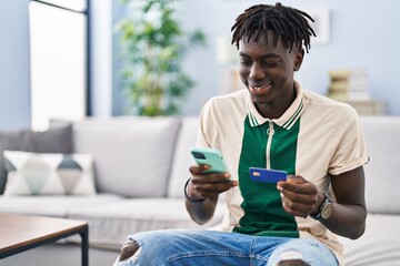 African american man using smartphone and credit card sitting on sofa at home