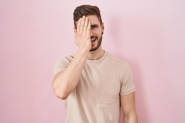 Hispanic man with beard standing over pink background covering one eye with hand, confident smile on face and surprise emotion.