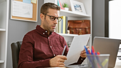 Fototapeta premium Concentrated hispanic man with glasses reviewing documents in a modern office
