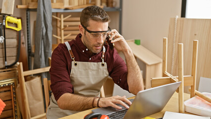 Handsome hispanic man with beard wearing glasses and apron uses laptop and phone in carpentry workshop. - obrazy, fototapety, plakaty