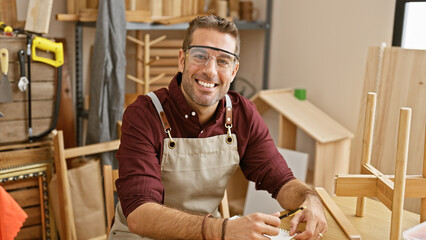 Handsome hispanic man with a beard wearing safety goggles smiles in a woodworking workshop.