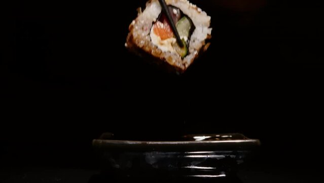 Hand using chopsticks dipping sushi roll into soy sauce in gravy boat on black background close-up.