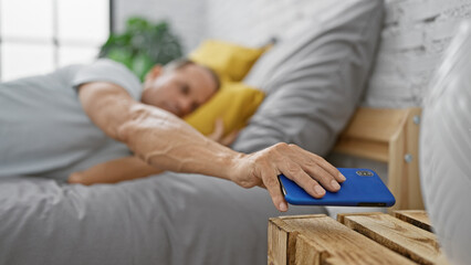 Relaxed hispanic middle age man happily waking up, holding his mobile phone comfortably resting in...