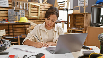 Head-turning young hispanic woman carpenter, meticulously taking notes on laptop in a vibrant carpentry workshop, embracing her woodworking business with professionalism