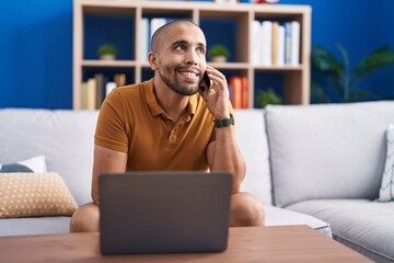Fototapeta premium Young latin man talking on smartphone using laptop at home