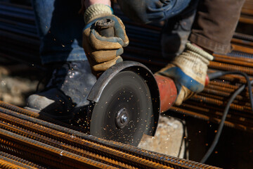 A builder in protective gloves holds a tool circular saw and cuts metal rods at a construction site