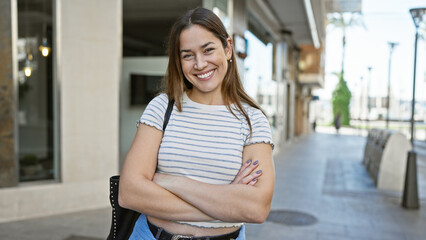 Fototapeta premium A smiling young woman with long brunette hair and blue eyes stands outdoors in a city street, arms crossed.