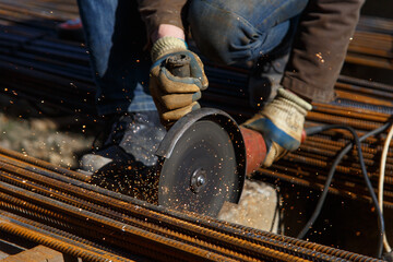 A builder in protective gloves holds a tool circular saw and cuts metal rods at a construction site