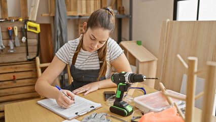 Focused woman scribbles notes in a woodshop brimming with tools, exuding a diy spirit amidst...