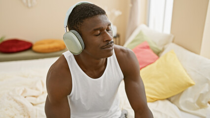 A relaxed african man chilling indoors on a bed, enjoying music with wireless headphones in a cozy...