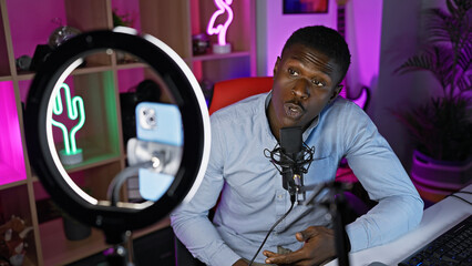 African american man podcasting with microphone and ring light in a colorful gaming room at home.