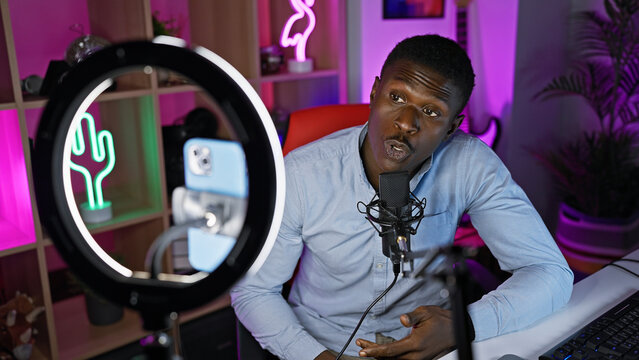 African american man podcasting with microphone and ring light in a colorful gaming room at home. - Powered by Adobe