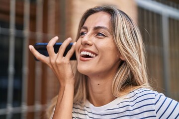 Young blonde woman miling confident listening audio message by the smartphone at street