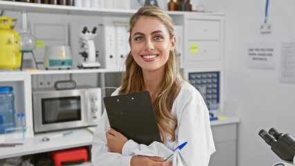 Confident blonde woman scientist masterfully balancing work and cheer, standing in a buzzing lab, smiling wide while effortlessly holding her clipboard amidst high-tech medical analysis.