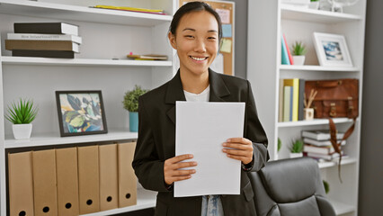 A professional chinese woman smiling, holding documents in a modern office setting with shelving...
