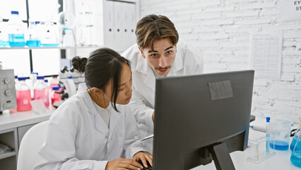 A man and woman work together in a laboratory, looking intently at a computer screen amidst scientific equipment.