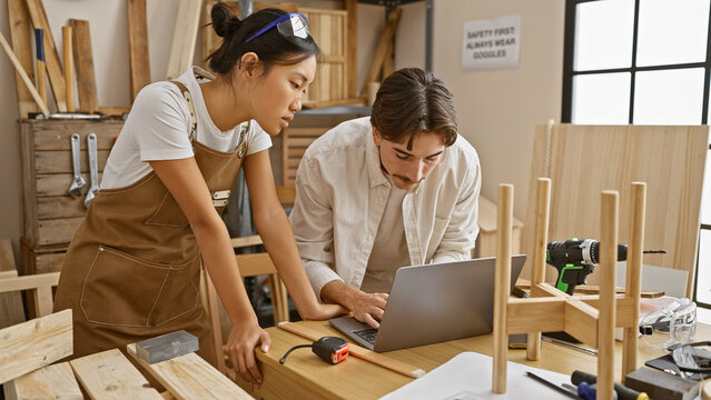 A man and woman craftsmen collaborating in a woodworking workshop while consulting a laptop.