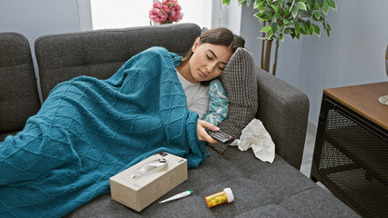 A young woman rests on a couch under a blue blanket, holding a remote, surrounded by tissues, a...