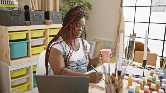 Beautiful african american woman artist painting a portrait, drawing on notebook whilst delightfully drinking morning coffee in her cozy art studio