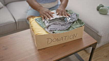 Warm-hearted african american woman putting folded clothes into a cardboard box for charity...